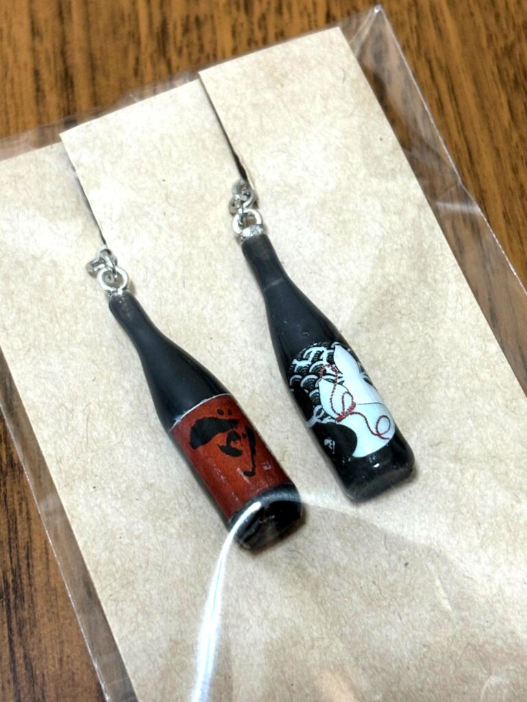 Earrings shaped like sake bottles, featuring labels of “Saki” Junmai Daiginjo and “monochrome+”