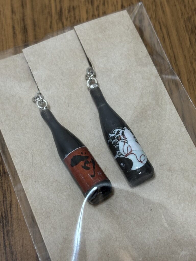 Earrings shaped like sake bottles, featuring labels of “Saki” Junmai Daiginjo and “monochrome+”