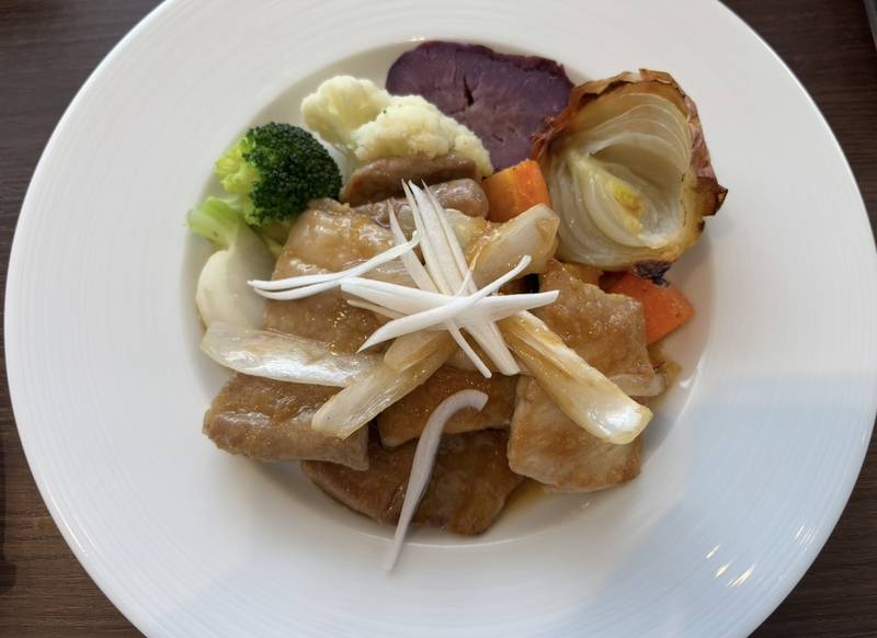 A plate of ginger-flavored pork steak served on a white dish, accompanied by roasted onion, broccoli, cauliflower, purple sweet potato, carrot, and other vegetables.