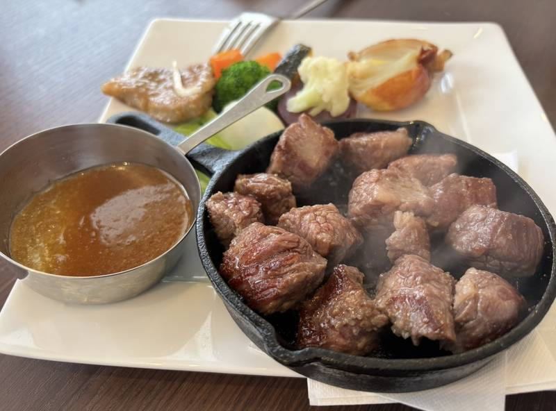 Sizzling wagyu dice steak served in a hot skillet, with a special sauce in a stainless steel cup and an assortment of vegetables on the side.