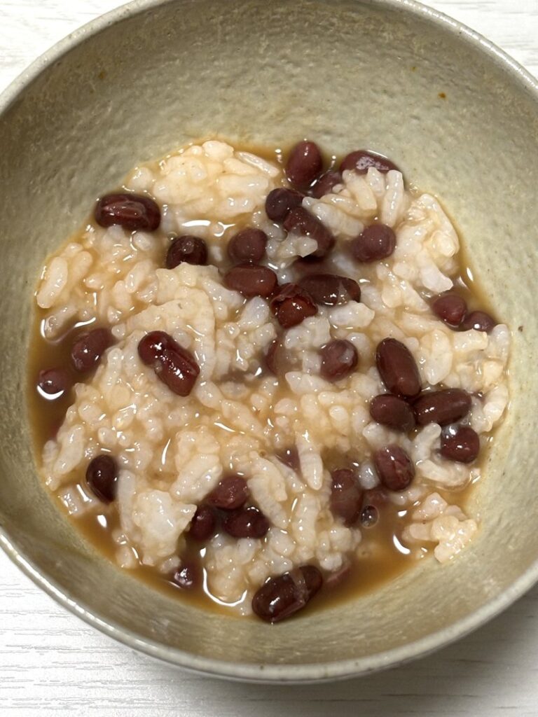 A bowl of savory azuki bean porridge with soft rice and red beans in a gentle, amber-colored broth.