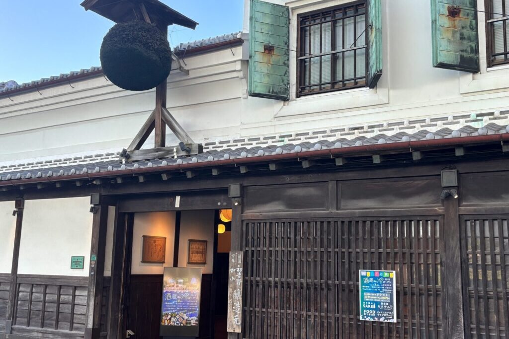 Front entrance of Hakata Hyakunen-gura sake brewery, featuring traditional white plaster walls, wooden lattice, verdigris shutters, and a sugidama.
