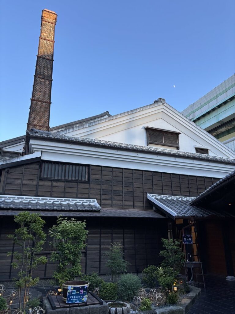 Exterior of Hakata Hyakunen-gura under a blue sky, featuring wooden walls, white plaster storehouse, and a tall brick chimney.
