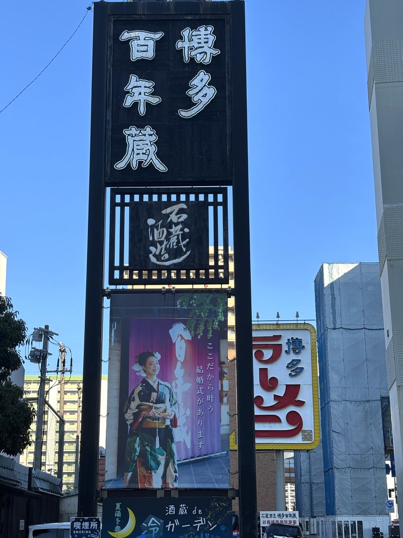 Signboard of Hakata Hyakunen-gura with a wedding advertisement below, set against a clear blue sky.