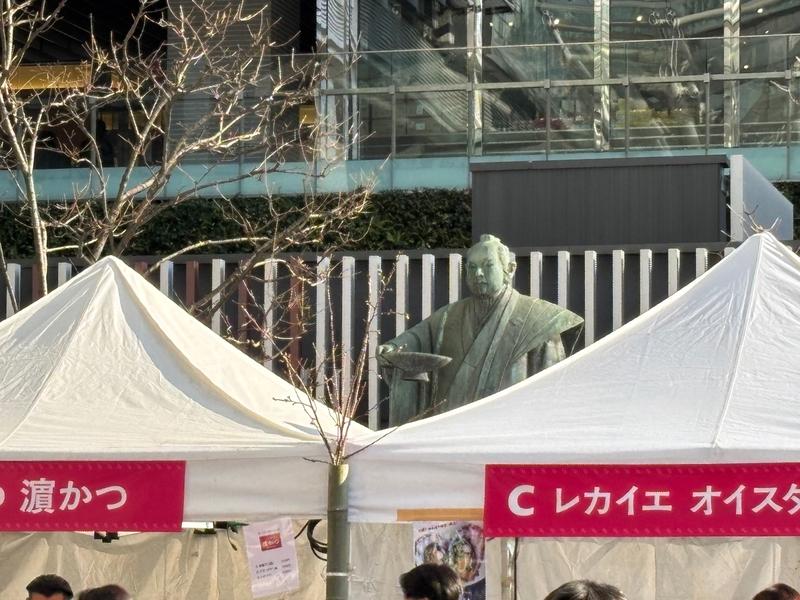 Behind the white tents, the Kuroda-bushi statue in front of Hakata Station is visible. The event venue is crowded with many people.