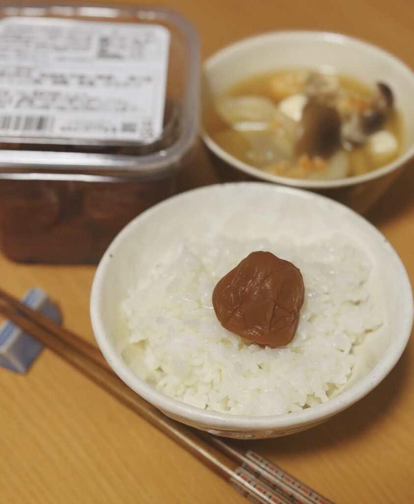 A bowl of white rice topped with a single Kishu Baikou umeboshi. In the background, a bowl of miso soup and a pack of umeboshi are visible.