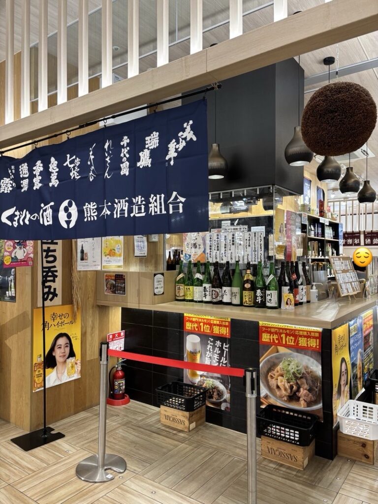 The standing bar corner of Ushijima Saketen inside Higo Yokamon Ichiba at Kumamoto Station. A noren banner of the Kumamoto Sake Brewers Association and a counter lined with local sake bottles.