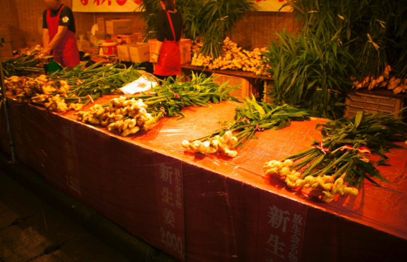 Bundles of fresh ginger lined up at a Hōjōya festival stall, on a red-covered table with vendors in the background.