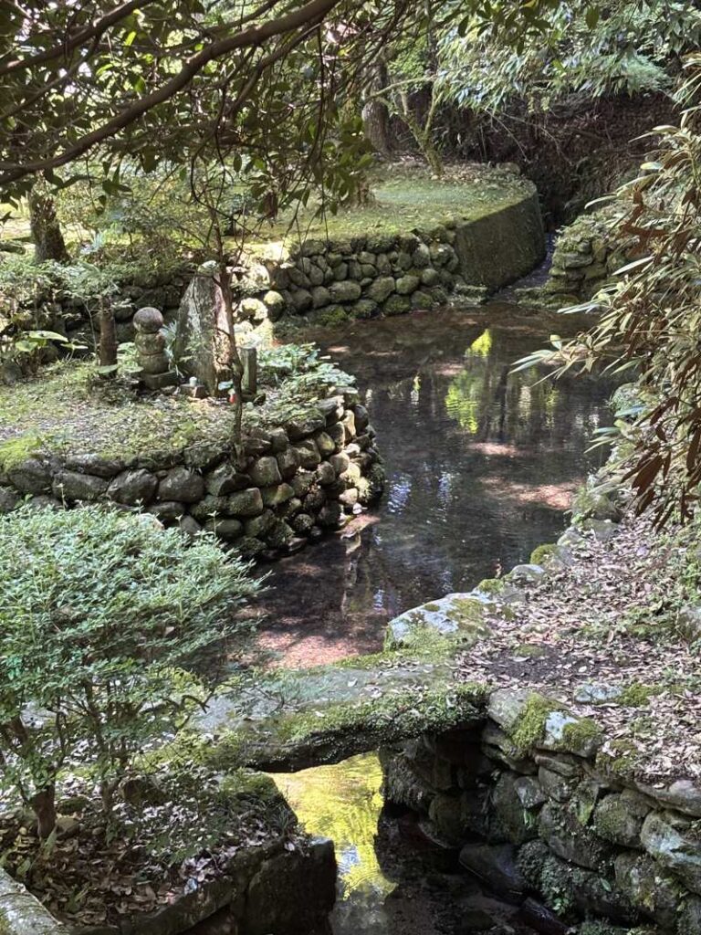 A clear spring pool at Shimizu Spring, ringed by stone walls and a small mossy footbridge with dappled light on the water.