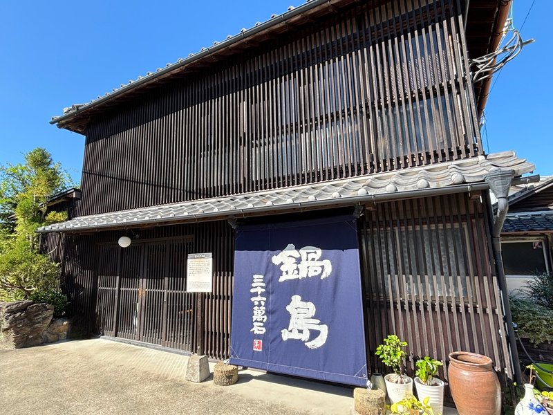 Exterior of Fukuchiyo Sake Brewery, featuring a wooden lattice facade and a navy noren curtain with the word “Nabeshima.”