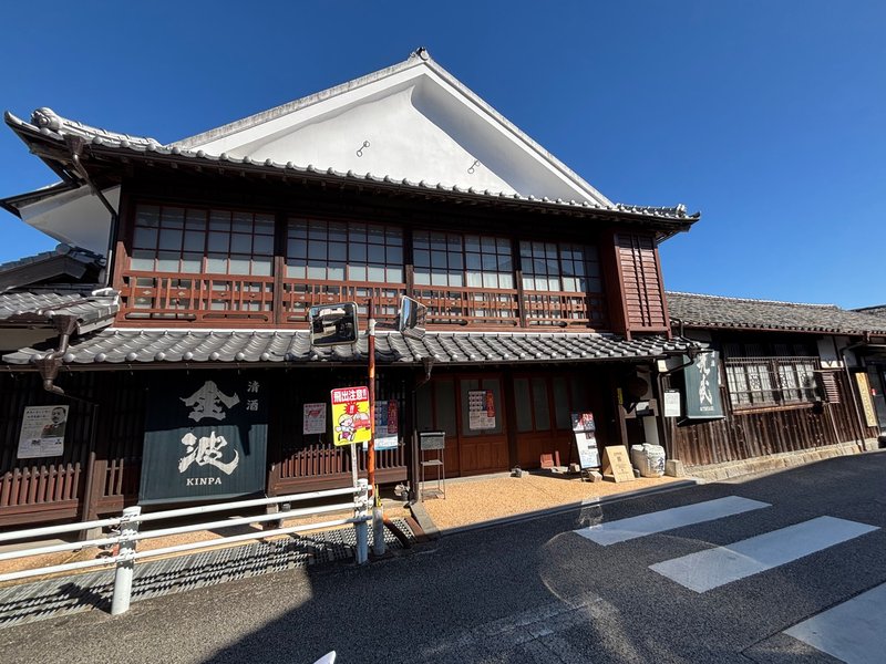 Exterior of Mitsutake Sake Brewery, featuring white plaster walls and wooden latticework with a noren curtain displaying the brand “Kinpa.”