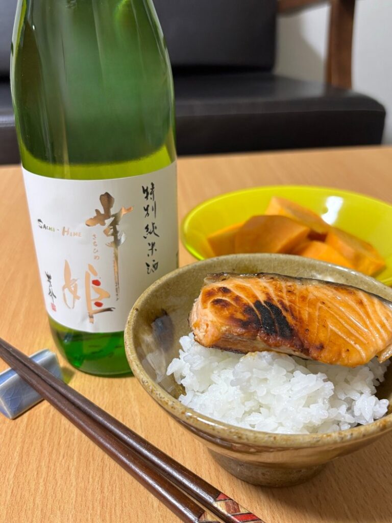 A bowl of rice topped with grilled salmon, served with persimmons in the background and a bottle of Tokubetsu Junmai Sake “Sachi-Hime.”