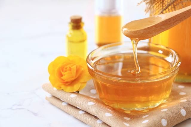 A photo of golden honey being poured into a glass bowl with a wooden spoon.