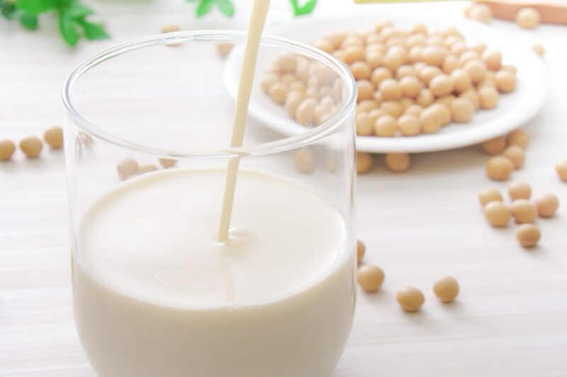 A glass of soy milk being poured, with a plate of soybeans in the background.
