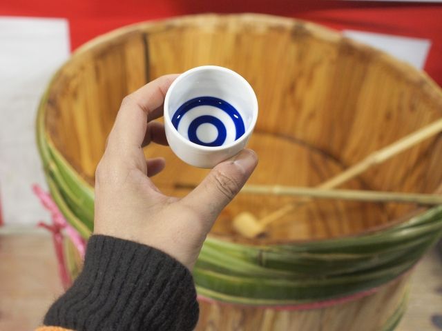 English: A hand holding a small cup of taruzake in front of a wooden sake barrel at a brewery event