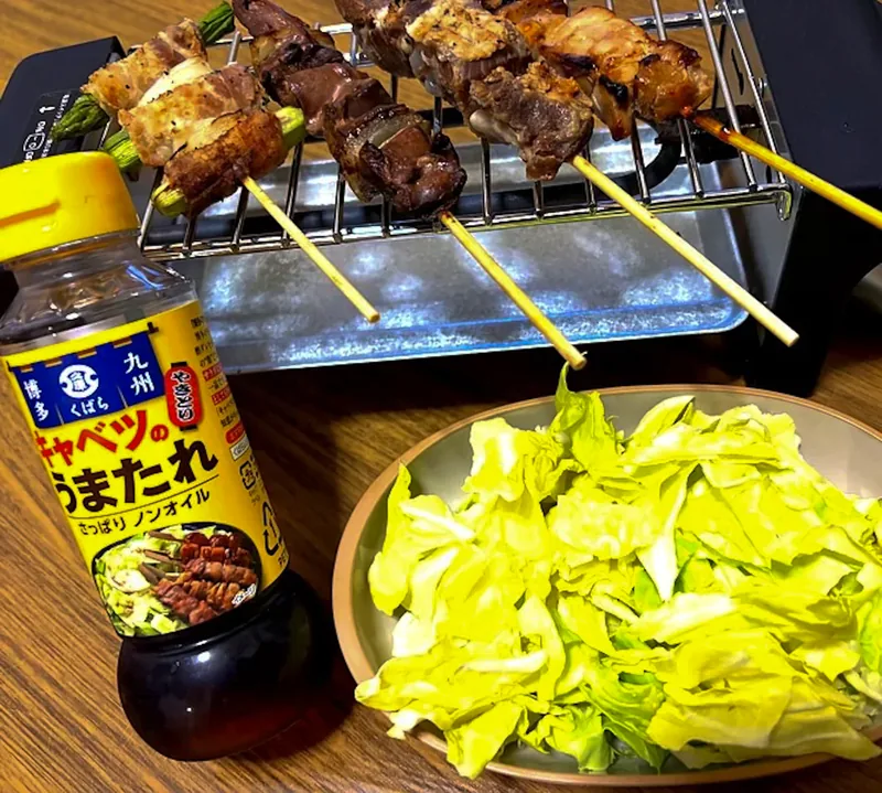A home yakitori set featuring grilled chicken skewers on a tabletop grill, a bottle of "Cabbage Uma-tare" sauce, and a plate of fresh shredded cabbage