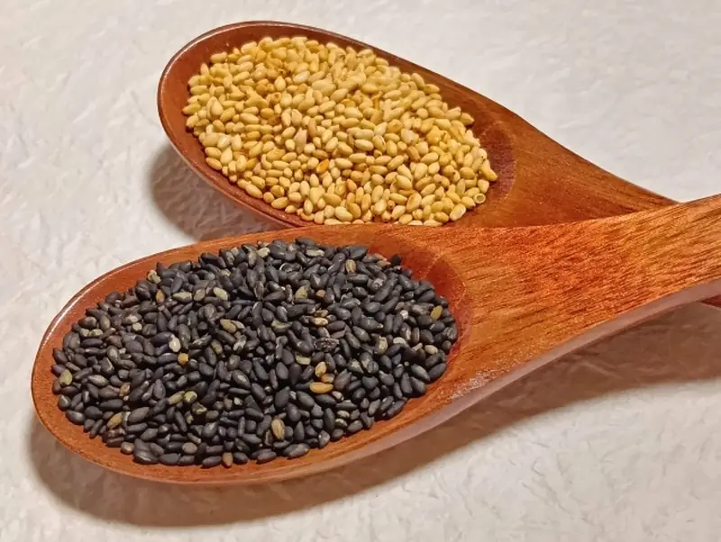 Two wooden spoons side by side, one holding golden sesame seeds and the other holding black sesame seeds