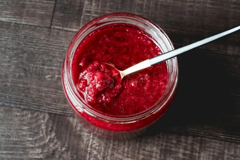 A glass jar of vibrant deep-red mulberry jam with a spoon resting on top