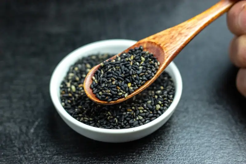 Black sesame seeds in a white bowl, scooped with a wooden spoon