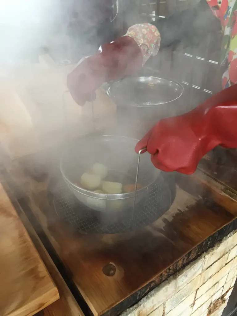 A person wearing red gloves handles a basket holding ingredients above a steaming jigoku-mushi cooking pit.