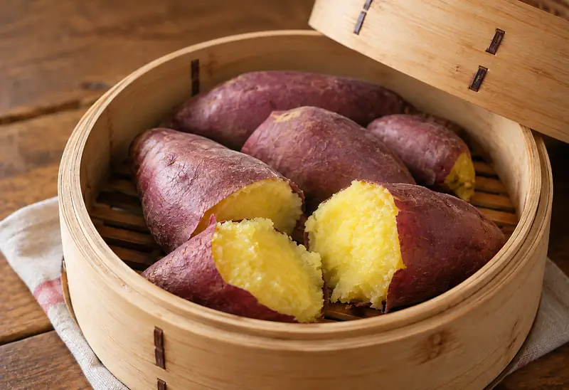 Skin-on steamed sweet potatoes in a bamboo steamer, with broken pieces in front revealing their golden flesh.